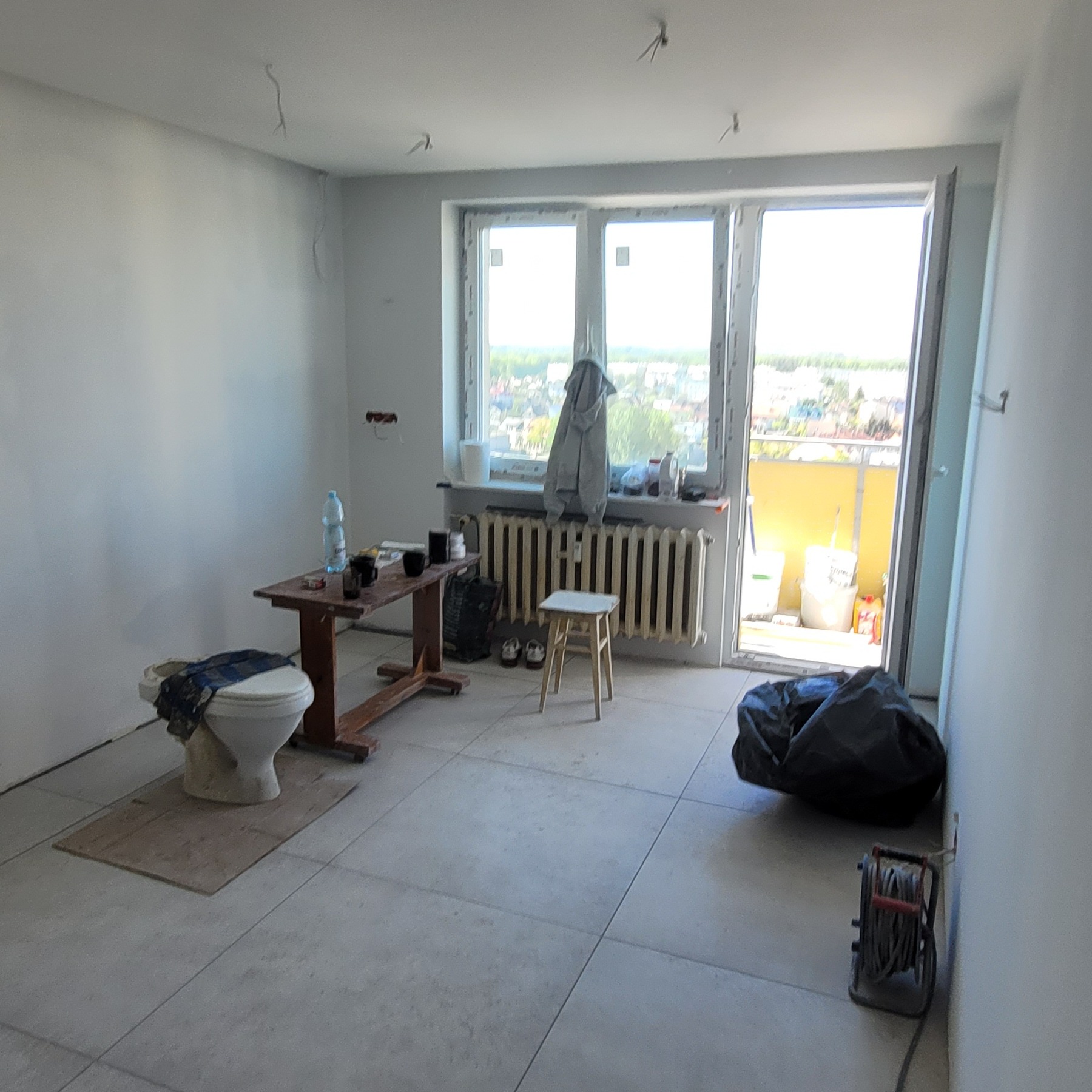 Wnętrze pokoju w trakcie remontu; widoczne gładkie, białe ściany, szare płytki podłogowe, prowizoryczny stolik z napojami, zakryta toaleta, grzejnik pod oknem balkonowym i zwisające kable z sufitu.