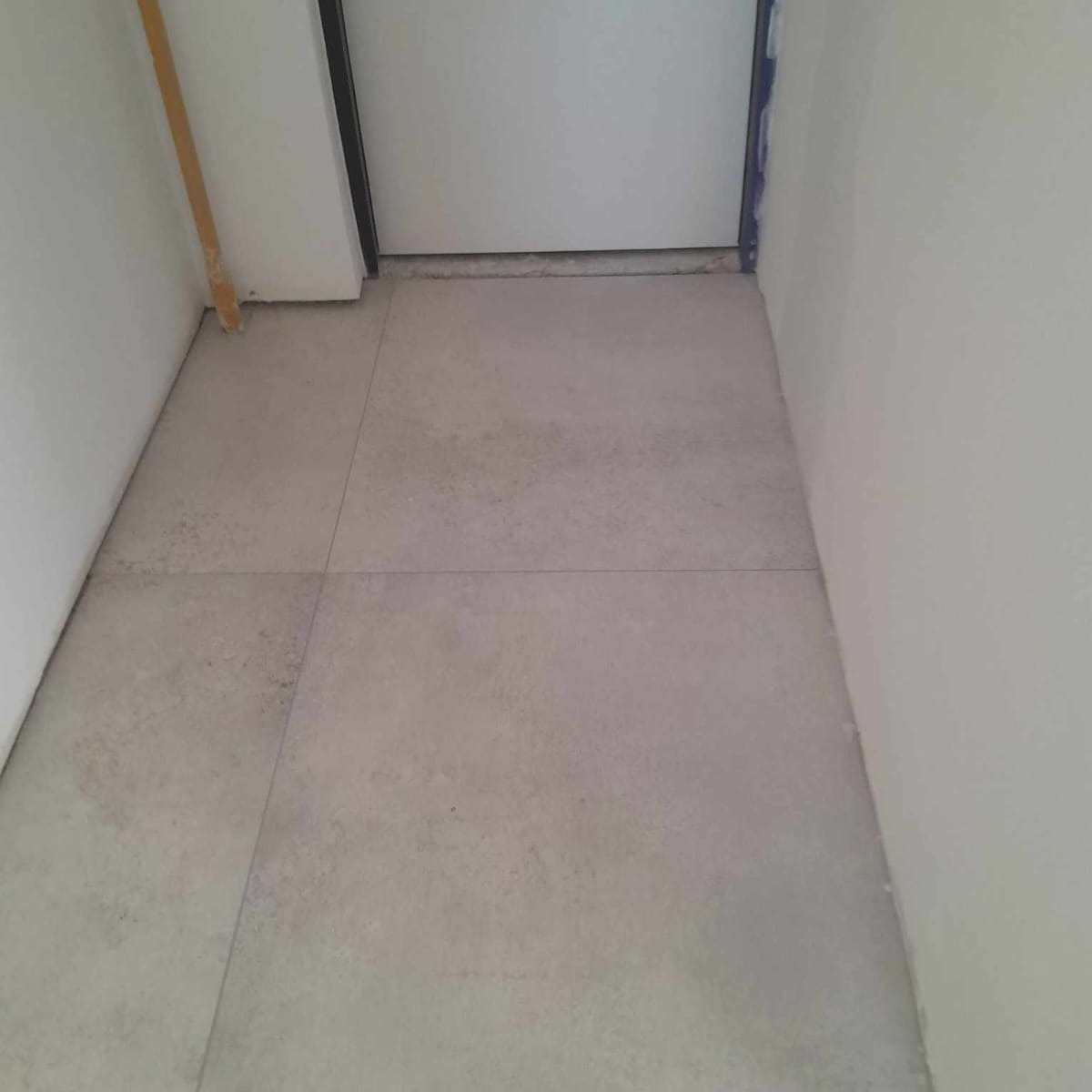 Wykończona podłoga wyłożona jasnoszarymi, matowymi płytkami w wąskim przedpokoju; widoczna biała ściana i drzwi wejściowe.