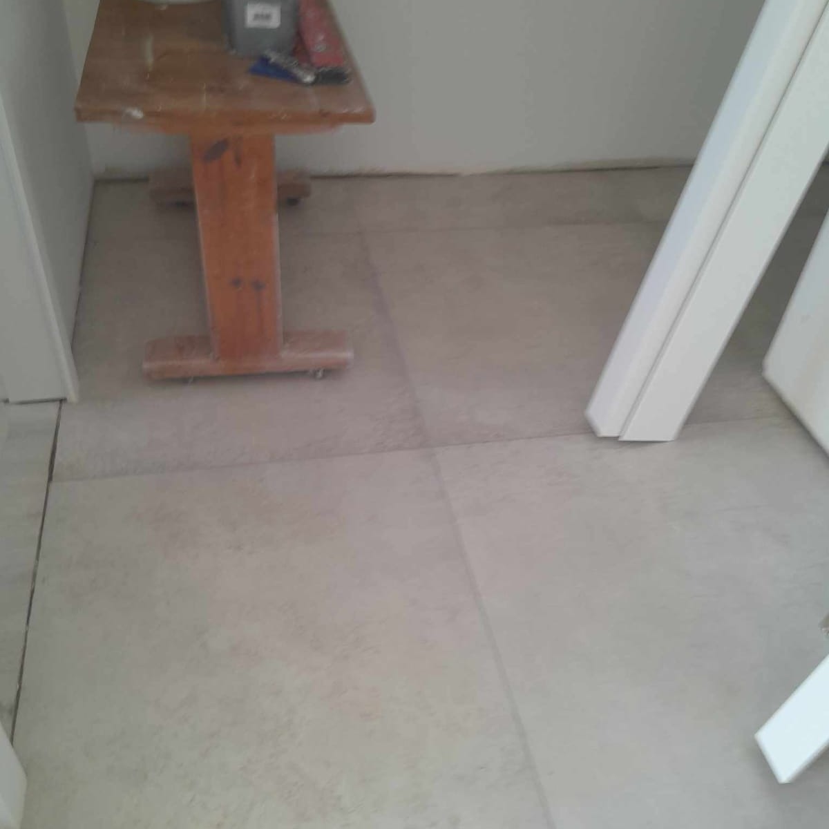 Ułożone jasnoszare płytki podłogowe z widocznym drewnianym stołem roboczym i fragmentem białej framugi drzwiowej.