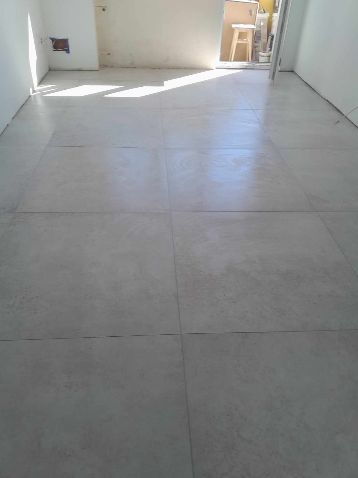 Ułożona podłoga z dużych, matowych, szarych płytek w nowym, jasnym pomieszczeniu, z widocznym fragmentem ściany z otworem na instalację elektryczną i fragmentem krzesła przez otwarte drzwi.