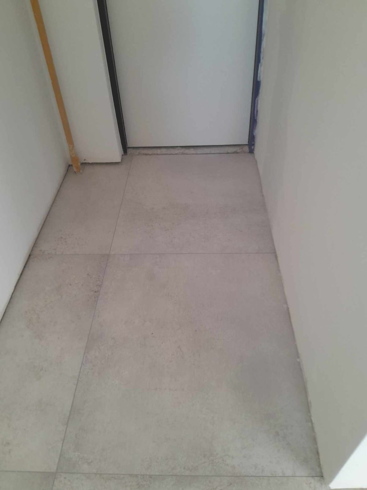 Ułożona podłoga z szarych, matowych płytek w korytarzu, widoczna biała ściana z listwą przypodłogową i fragment ramy drzwi.