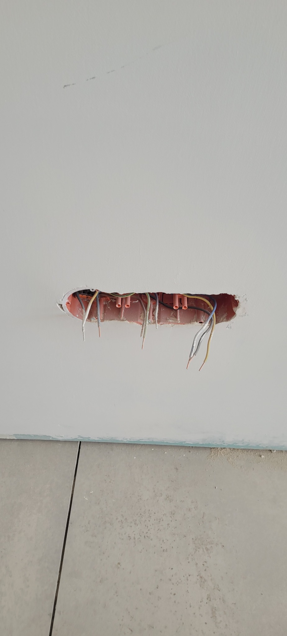 Otwór w suficie z wystającymi przewodami elektrycznymi, widoczne rurki instalacyjne w kolorze pomarańczowym, surowy stan wykończenia, płytki podłogowe w tle.