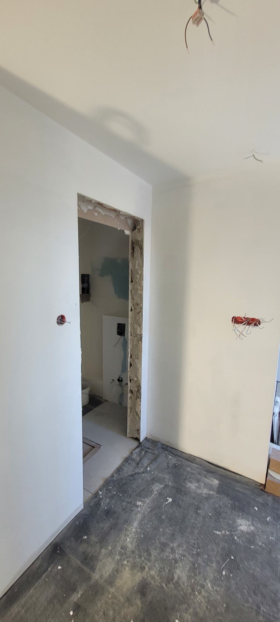 Pokój w trakcie remontu: ściany po szpachlowaniu i malowaniu na biało, widoczne przewody elektryczne wystające ze ściany i sufitu, surowa ościeżnica drzwiowa, podłoga zabezpieczona folią.