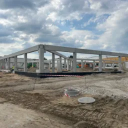 Konstrukcja szkieletowa budynku w trakcie budowy, widoczne betonowe słupy i belki, plac budowy z ciężkim sprzętem, ziemia z wykopami, w tle błękitne niebo z chmurami.