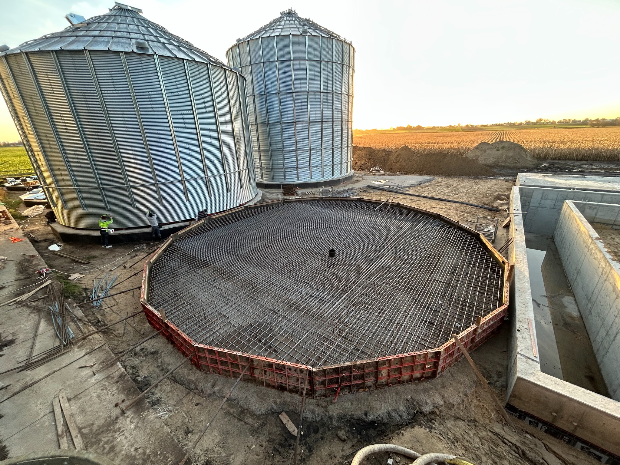 Zbrojenie okrągłego fundamentu pod silos zbożowy, widok z góry. Stalowa siatka zbrojeniowa w szalunku, w tle dwa silosy i pole uprawne oświetlone zachodzącym słońcem.