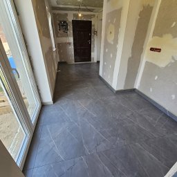 Michael Berg - Wnętrze w trakcie prac wykończeniowych: szare płytki podłogowe z białym żyłkowaniem, ściany przygotowane pod malowanie z widocznymi łatami gipsowymi, widoczna instalacja elektryczna, drzwi...