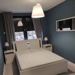 Michael Berg - Jasna sypialnia z białym łóżkiem, szafkami nocnymi i lampkami. Na ścianie niebieska farba, a okno zasłonięte szarymi zasłonami i roletami. Widoczne białe drzwi z czarną klamką.