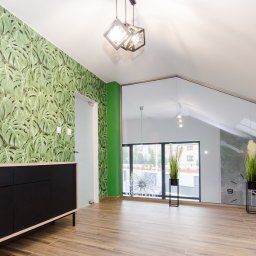 Michael Berg - Wnętrze z tapetą w liście monstery, czarną komodą i drewnopodobną podłogą, oświetlone nowoczesnymi lampami wiszącymi.