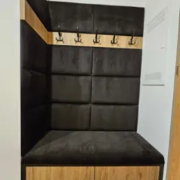 Wnęka z siedziskiem i wieszakami, tapicerowana ciemnoszarą tkaniną w geometryczne kwadraty, z drewnianymi elementami wykończeniowymi w kolorze jasnego drewna.