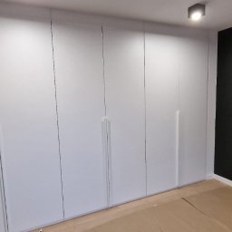 Michael Berg - Biała, minimalistyczna szafa wnękowa z długimi, pionowymi uchwytami na tle czarnej, tapicerowanej ściany z lampką nocną.