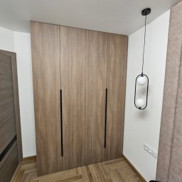 Michael Berg - Wysoka, zabudowana szafa z drewnopodobnymi frontami i długimi, czarnymi uchwytami, oświetlona wiszącą lampą z dwoma białymi kulami, na podłodze parkiet ułożony w jodełkę.