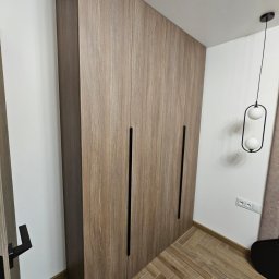 Michael Berg - Wysoka, zabudowana szafa z drewnopodobnymi frontami i długimi, pionowymi, czarnymi uchwytami, dopasowana do skosu sufitu, z widoczną lampą wiszącą z dwoma białymi kulami i parkietem ułożonym...