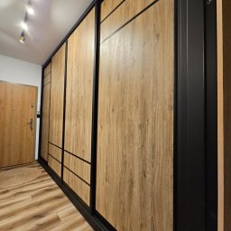 Michael Berg - Długa szafa wnękowa z przesuwnymi drzwiami o teksturze jasnego drewna, z czarnymi ramami i poziomymi akcentami, zamontowana w przedpokoju z drewnianą podłogą i widocznymi reflektorami sufitowymi.