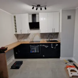 Nowoczesna kuchnia z czarnymi szafkami dolnymi i białymi górnymi, blat z jasnego drewna, widoczne narzędzia i akcesoria montażowe, butelka piwa na blacie, podłoga wyłożona płytkami.