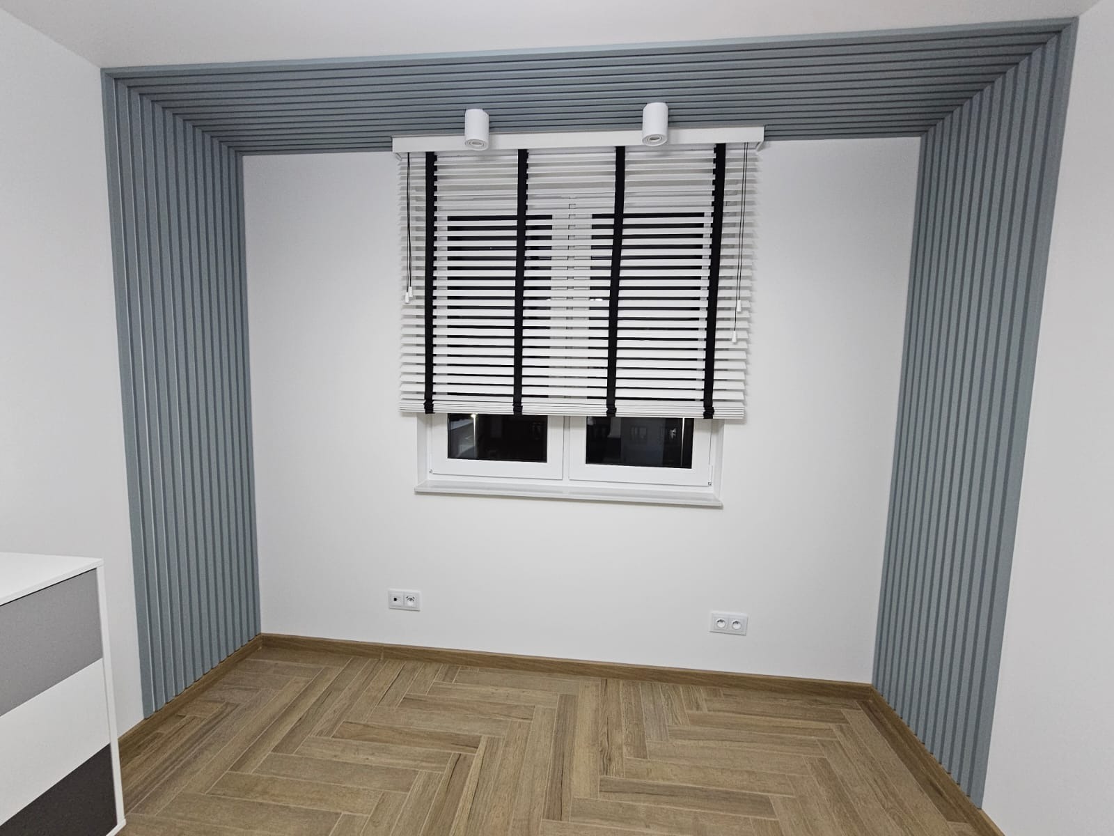 Pokój po remoncie: drewniana podłoga ułożona w jodełkę, białe ściany z szarymi panelami dekoracyjnymi na ścianach i suficie, okno z białymi żaluzjami z czarnymi taśmami.