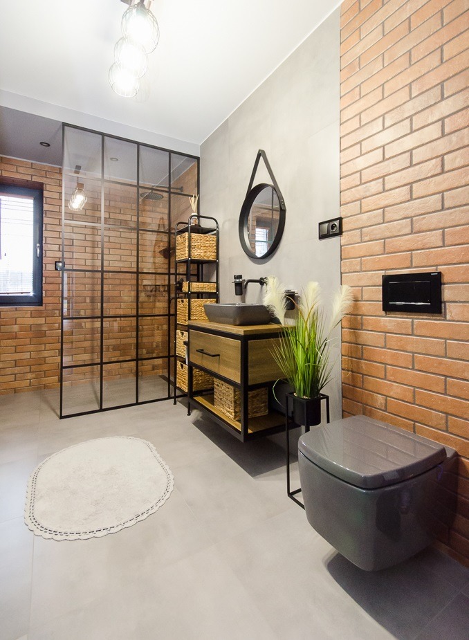 Nowoczesna łazienka z ceglaną ścianą, kabiną prysznicową typu walk-in z czarnymi szprosami, umywalką nablatową na drewnianej szafce z metalowym stelażem, okrągłym lustrem na skórzanym pasku...