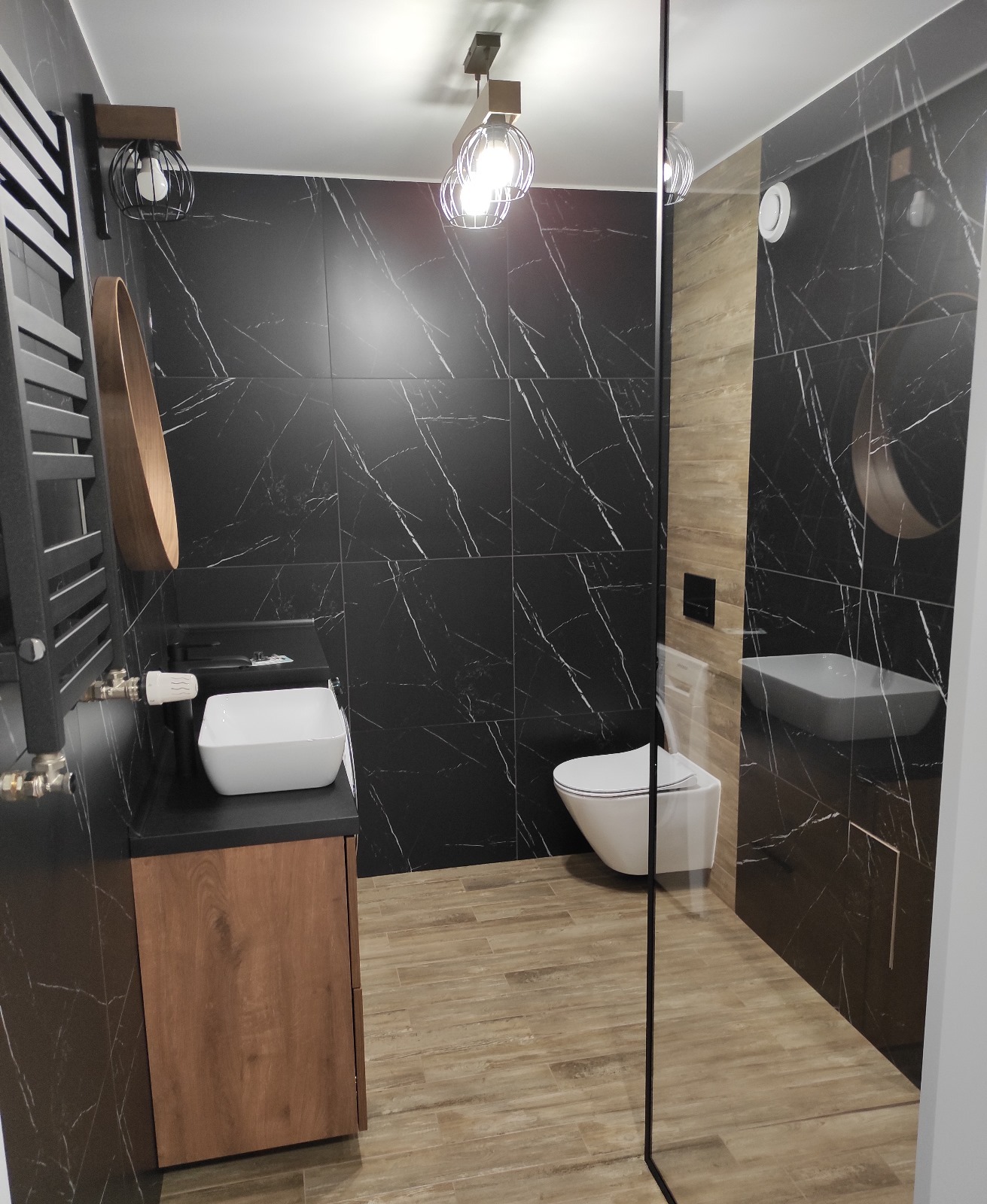 Nowoczesna łazienka z czarnymi płytkami imitującymi marmur, drewnianymi akcentami i minimalistyczną białą armaturą, oświetlona dekoracyjnymi lampami.