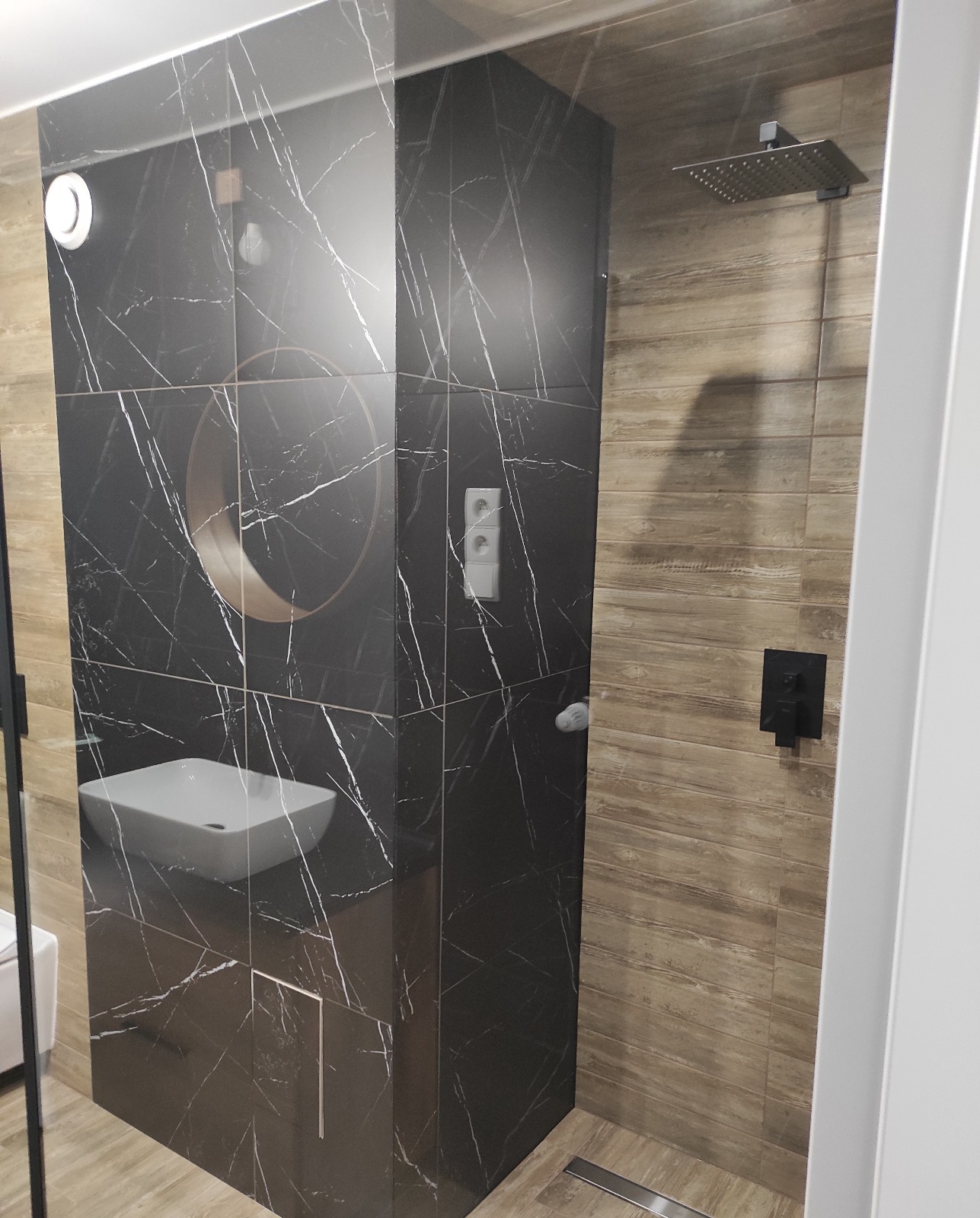 Nowoczesna łazienka z kabiną prysznicową typu walk-in, wykończona płytkami imitującymi marmur i drewno, z czarną armaturą i umywalką nablatową.