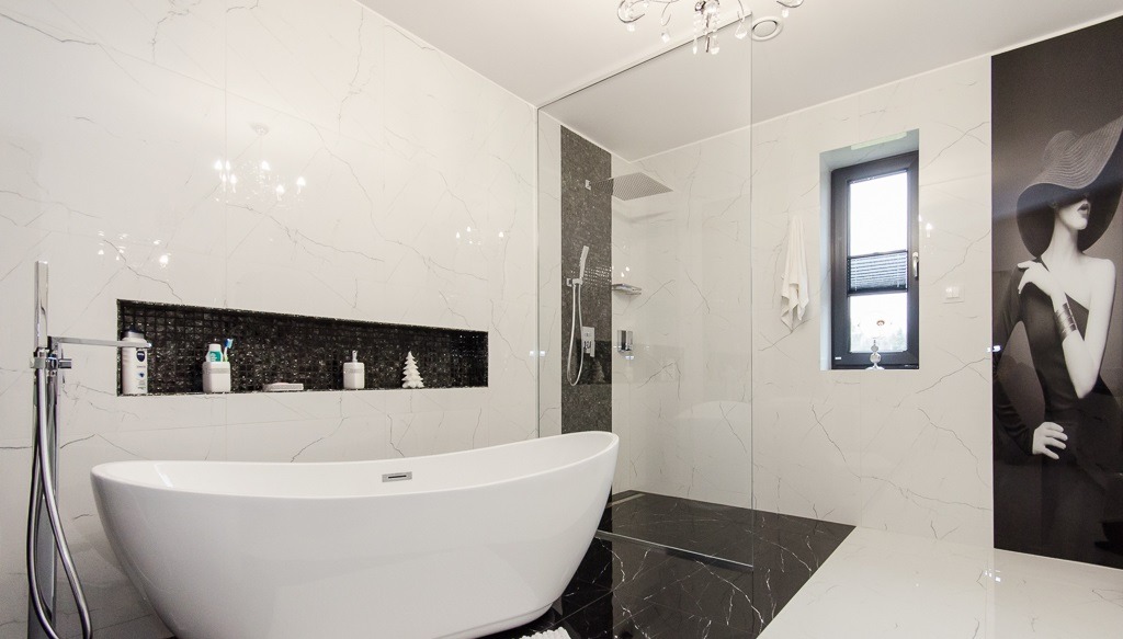 Elegancka łazienka z wolnostojącą białą wanną, czarno-białymi marmurkowymi płytkami, czarną mozaiką we wnęce i portretem kobiety w kapeluszu na ścianie.