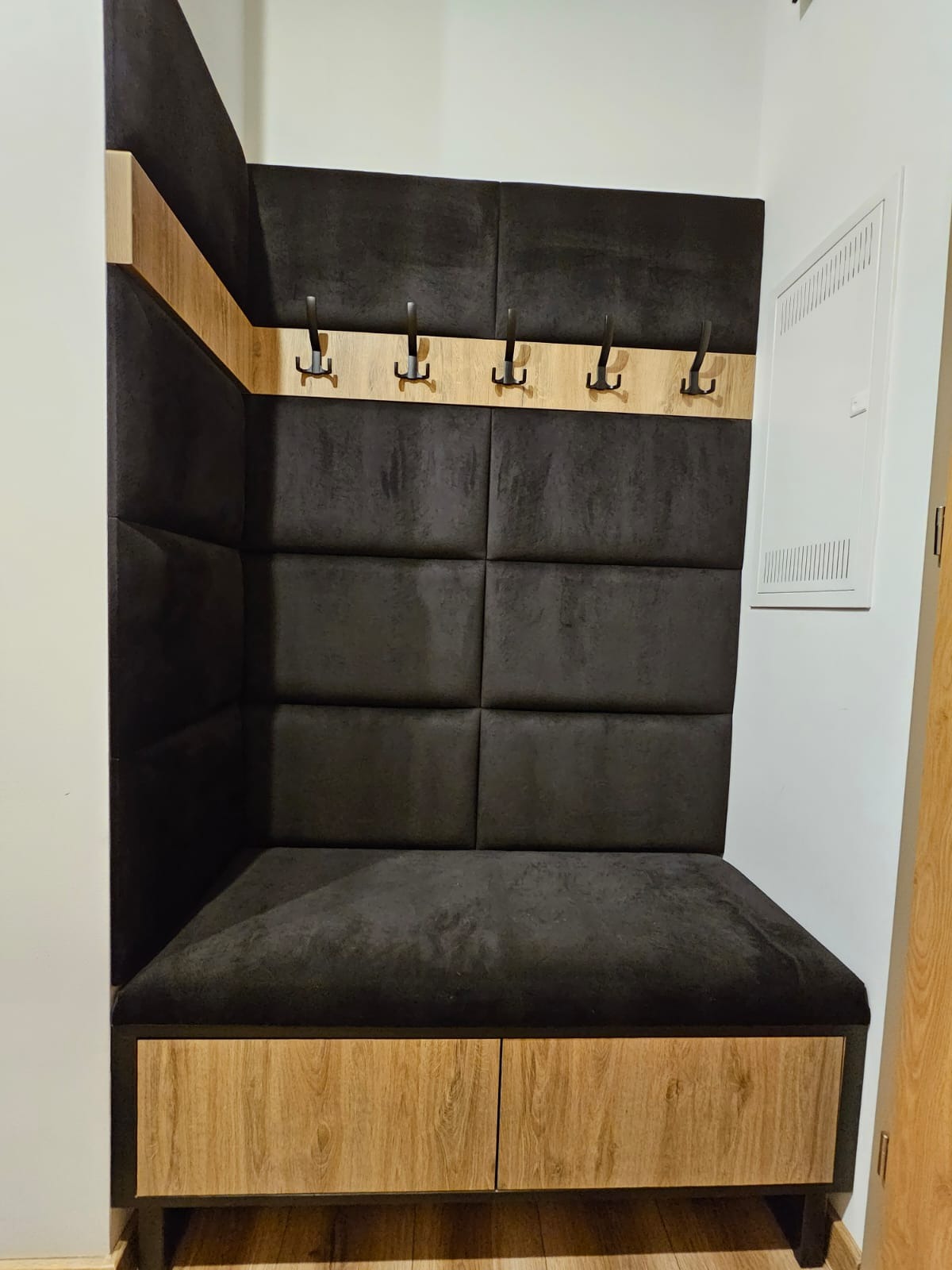 Wnęka z siedziskiem i wieszakami, tapicerowana ciemnoszarą tkaniną w geometryczne kwadraty, z drewnianymi elementami wykończeniowymi w kolorze jasnego drewna.