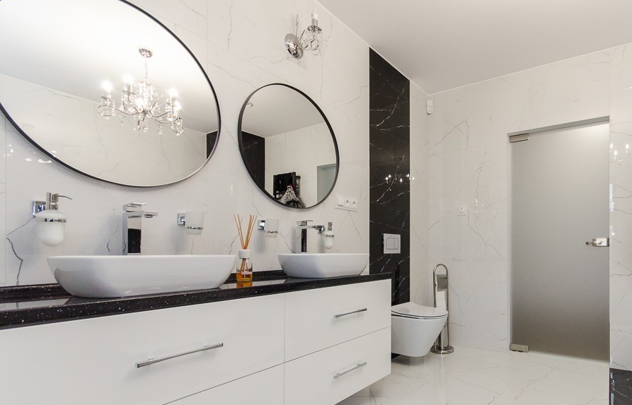 Nowoczesna łazienka z białymi marmurowymi płytkami, dwoma okrągłymi lustrami z czarną ramą, białymi umywalkami nablatowymi, czarnym blatem, białymi szafkami, toaletą wiszącą i szklanymi drzwiami...