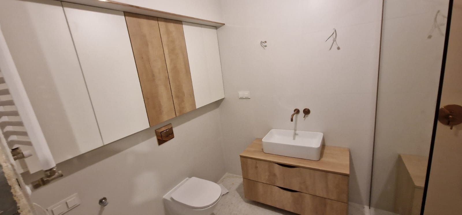 Nowoczesna łazienka w minimalistycznym stylu, z wiszącą szafką z białymi i drewnianymi frontami, białą umywalką nablatową z miedzianą armaturą i podwieszaną miską WC.