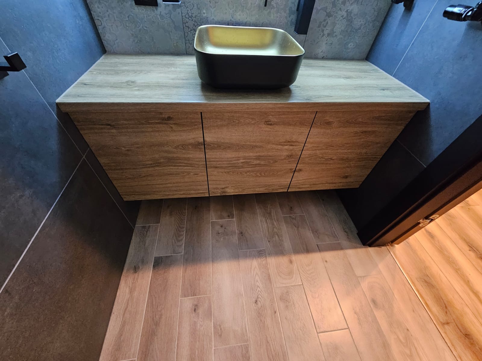 Nowoczesna łazienka z czarną umywalką o złotym wnętrzu, drewnopodobna szafka pod umywalką i podłoga z płytek imitujących drewno, ciemne ściany.