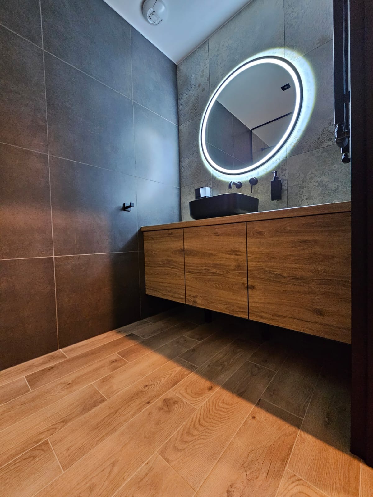 Nowoczesna łazienka z drewnianą szafką pod umywalkę, okrągłym lustrem z podświetleniem LED, czarną umywalką nablatową i grafitowymi płytkami na ścianach oraz drewnopodobną podłogą z płytek.