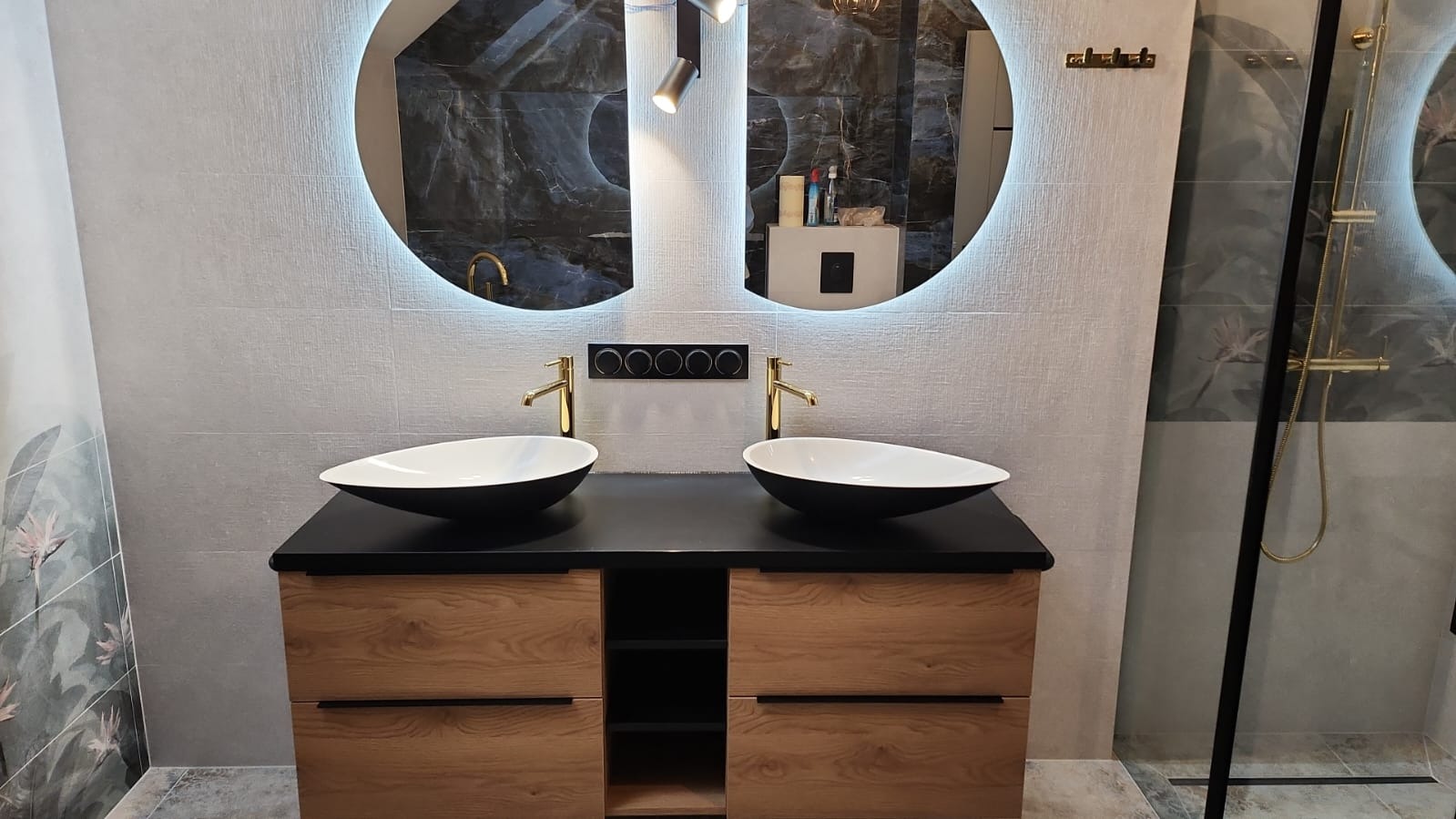 Nowoczesna łazienka z dwoma owalnymi umywalkami nablatowymi, złotą armaturą, podświetlanymi okrągłymi lustrami i kabiną prysznicową ze złotymi elementami.