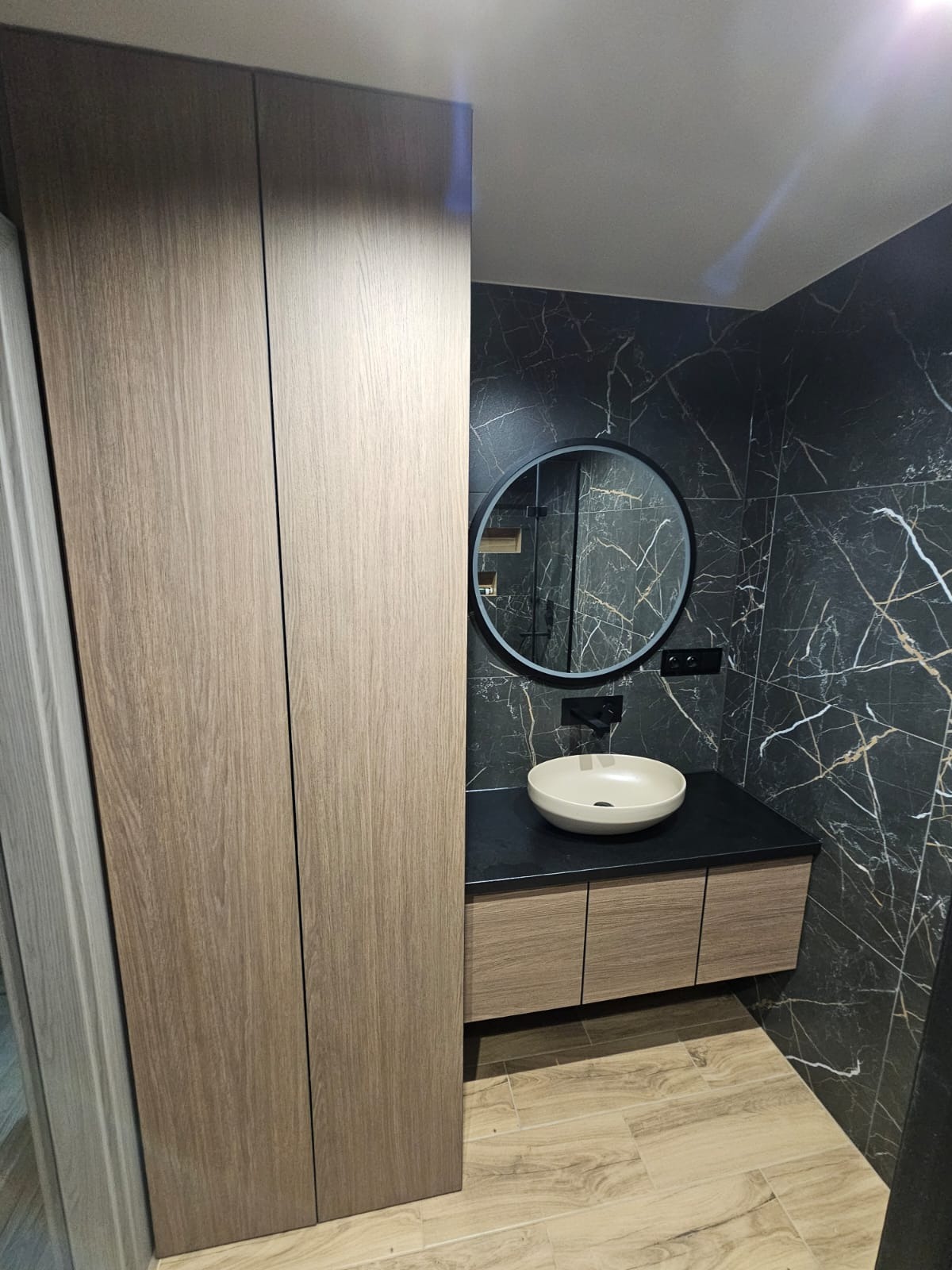 Nowoczesna łazienka z umywalką nablatową, okrągłym lustrem z czarną ramą, szafką pod umywalką z drewnopodobnymi frontami i wysoką szafą w zabudowie, na tle ściany wykończonej płytkami imitującymi...