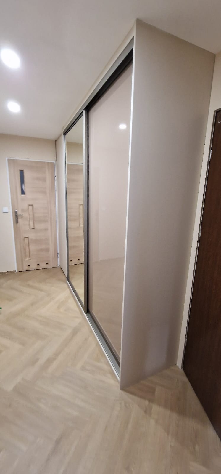 Beżowa szafa wnękowa z przesuwnymi drzwiami lustrzanymi, widok z boku na tle parkietu ułożonego w jodełkę.