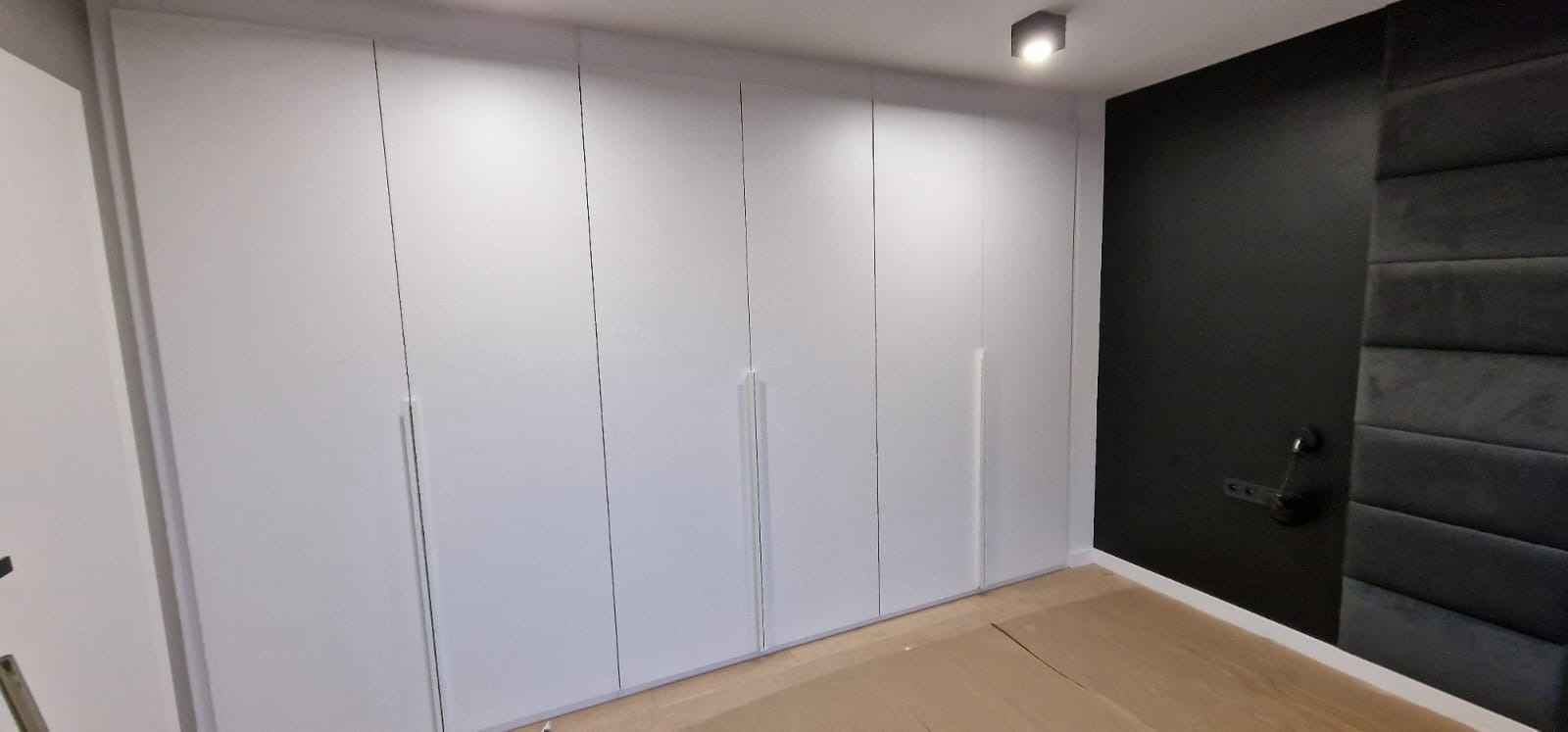 Biała, minimalistyczna szafa wnękowa z długimi, pionowymi uchwytami na tle czarnej, tapicerowanej ściany z lampką nocną.