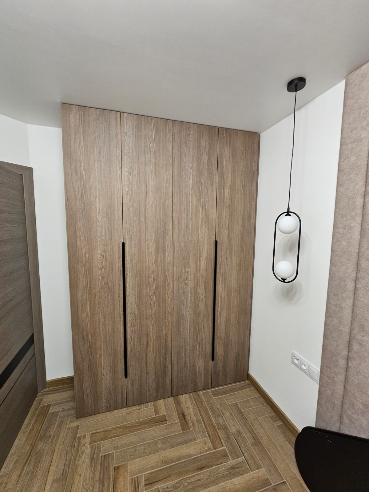 Wysoka, zabudowana szafa z drewnopodobnymi frontami i długimi, czarnymi uchwytami, oświetlona wiszącą lampą z dwoma białymi kulami, na podłodze parkiet ułożony w jodełkę.