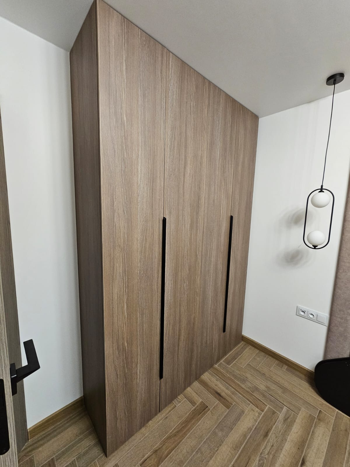 Wysoka, zabudowana szafa z drewnopodobnymi frontami i długimi, pionowymi, czarnymi uchwytami, dopasowana do skosu sufitu, z widoczną lampą wiszącą z dwoma białymi kulami i parkietem ułożonym...