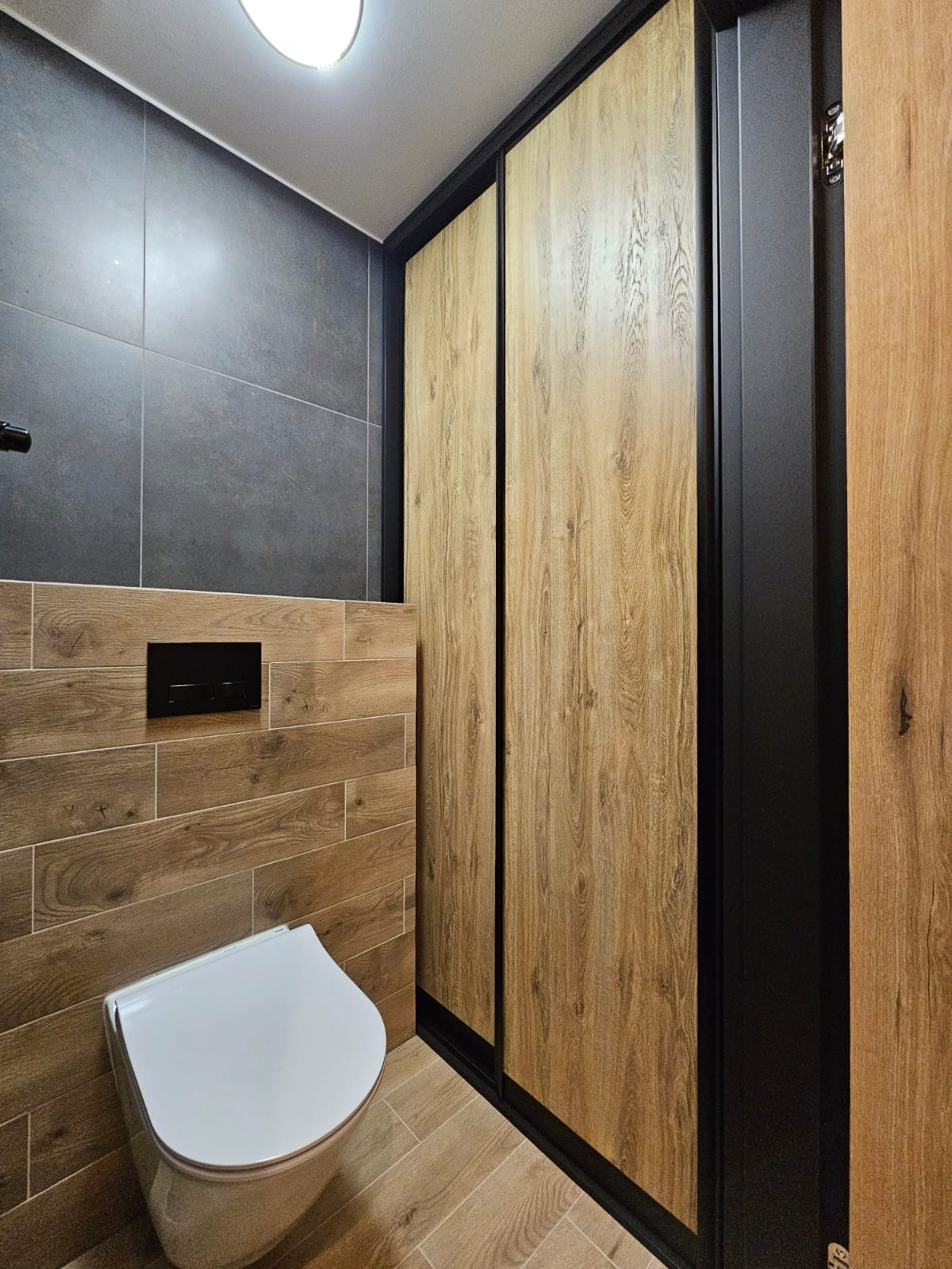 Nowoczesna łazienka z podwieszaną toaletą, brązowymi płytkami imitującymi drewno na ścianach i podłodze oraz przesuwnymi drzwiami szafy z okleiną drewnopodobną i czarnymi obramowaniami.