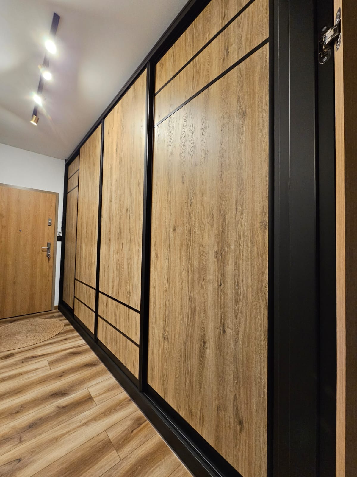 Długa szafa wnękowa z przesuwnymi drzwiami o teksturze jasnego drewna, z czarnymi ramami i poziomymi akcentami, zamontowana w przedpokoju z drewnianą podłogą i widocznymi reflektorami sufitowymi.