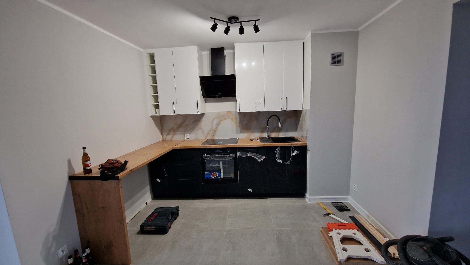 Nowoczesna kuchnia z czarnymi szafkami dolnymi i białymi górnymi, blat z jasnego drewna, widoczne narzędzia i akcesoria montażowe, butelka piwa na blacie, podłoga wyłożona płytkami.