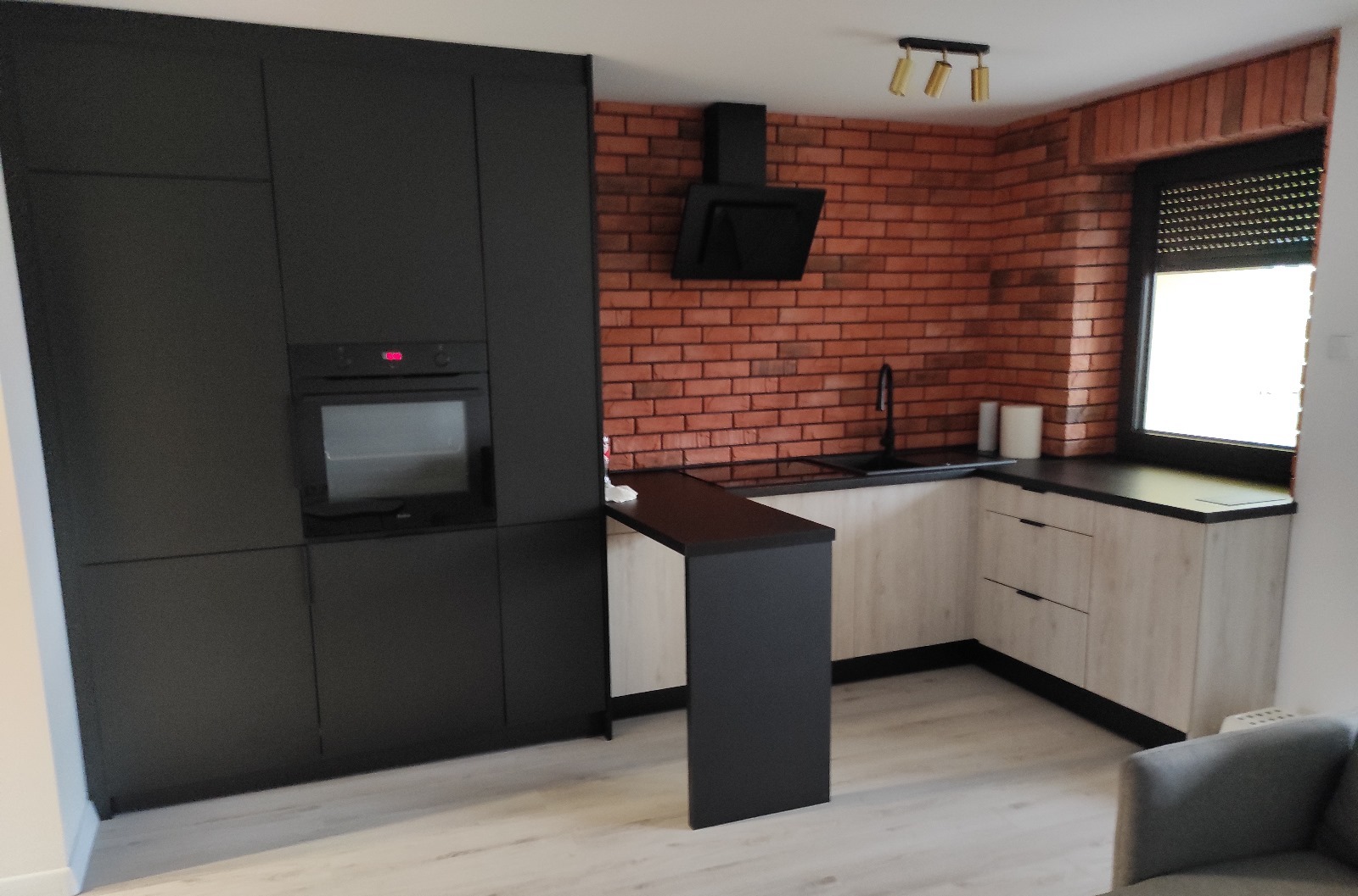 Nowoczesna kuchnia w Sosnowcu z czarnymi frontami szafek i ceglaną ścianą, oświetlona trzema lampami punktowymi, z blatem roboczym w kolorze czarnym.