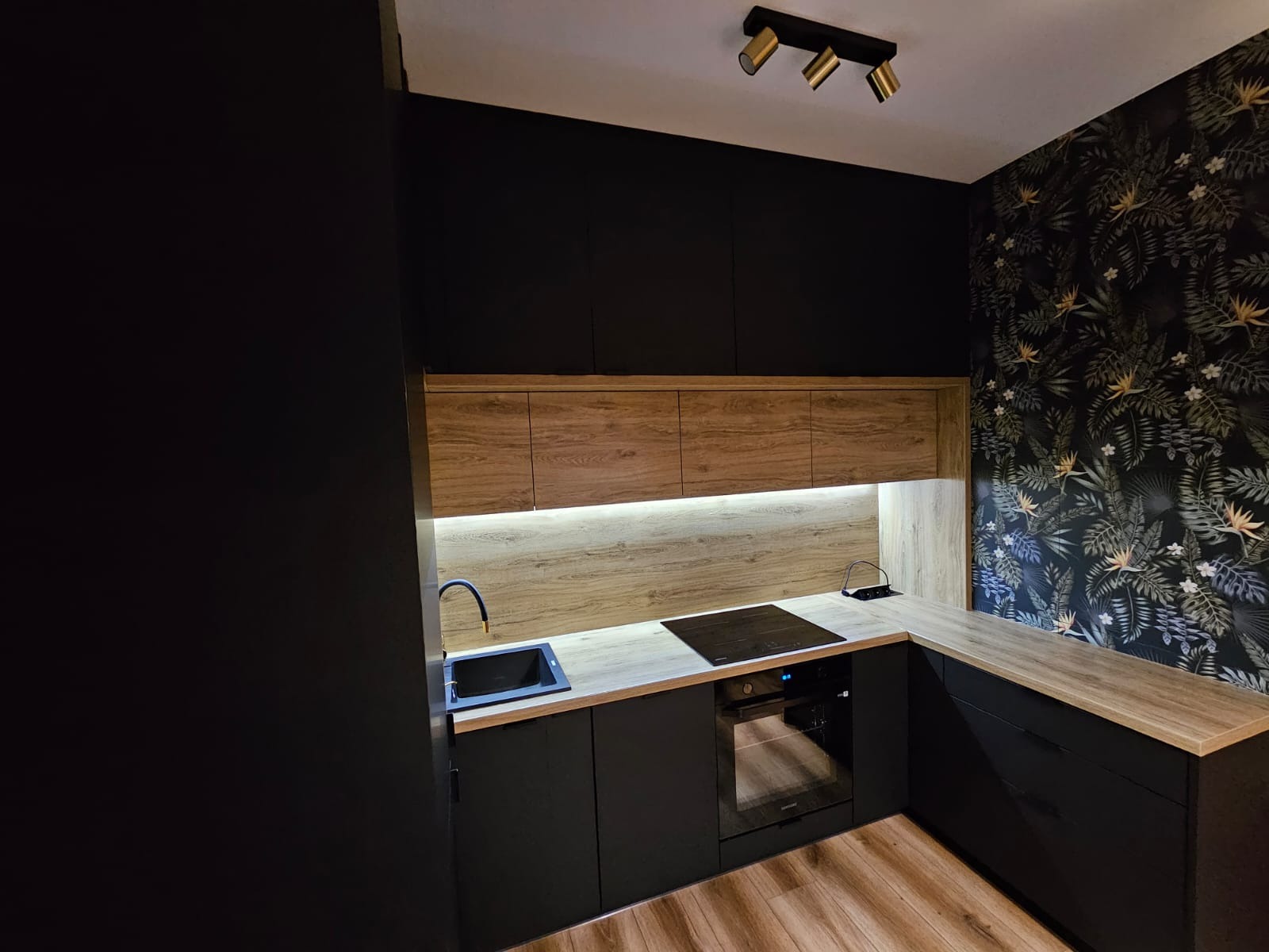 Nowoczesna kuchnia w czerni i drewnie z tropikalną tapetą, oświetleniem LED pod szafkami i minimalistycznym wyposażeniem.