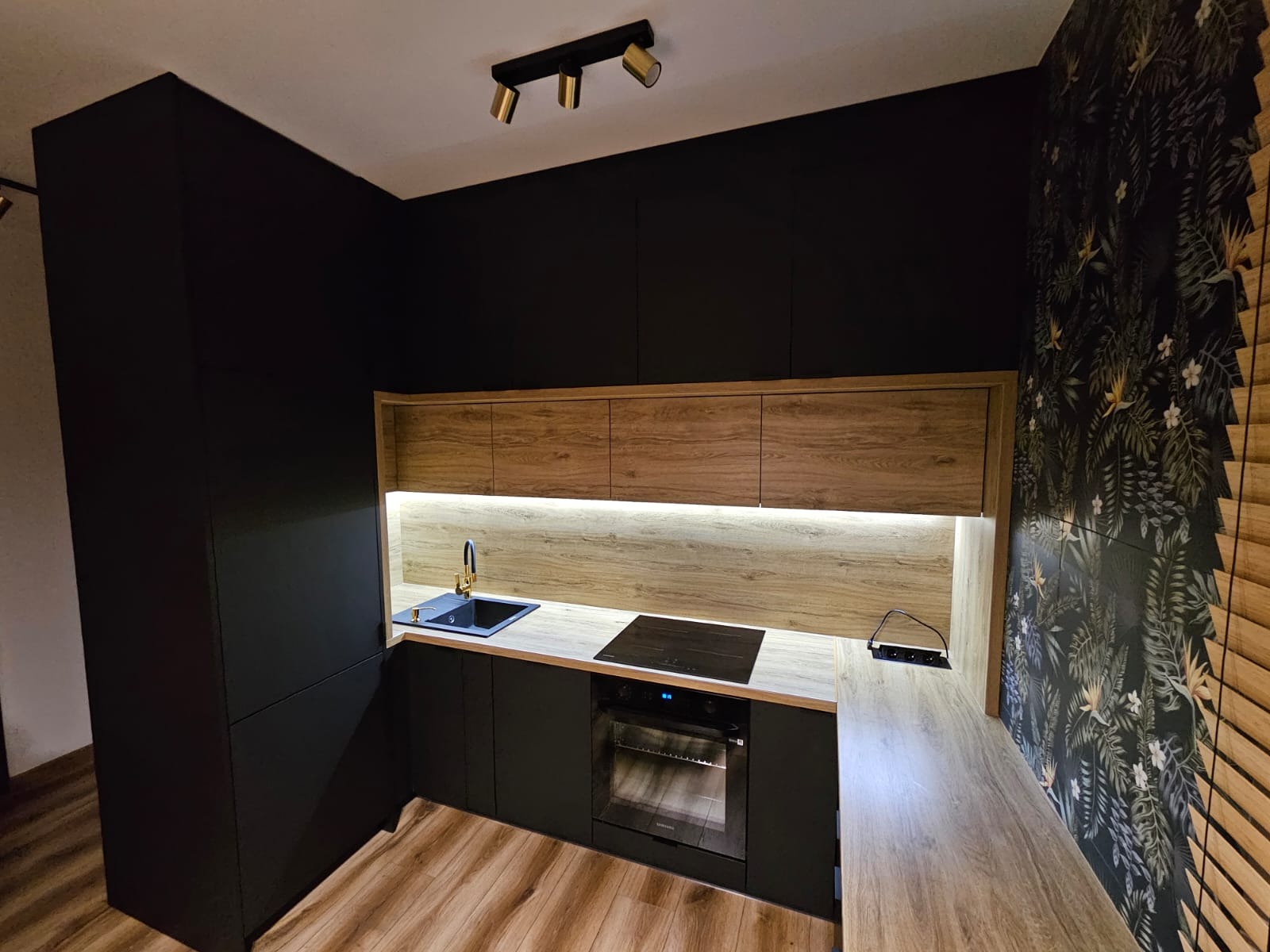 Nowoczesna kuchnia z czarnymi szafkami, drewnianymi frontami górnych szafek z oświetleniem LED, złotą armaturą i tapetą w tropikalne wzory.