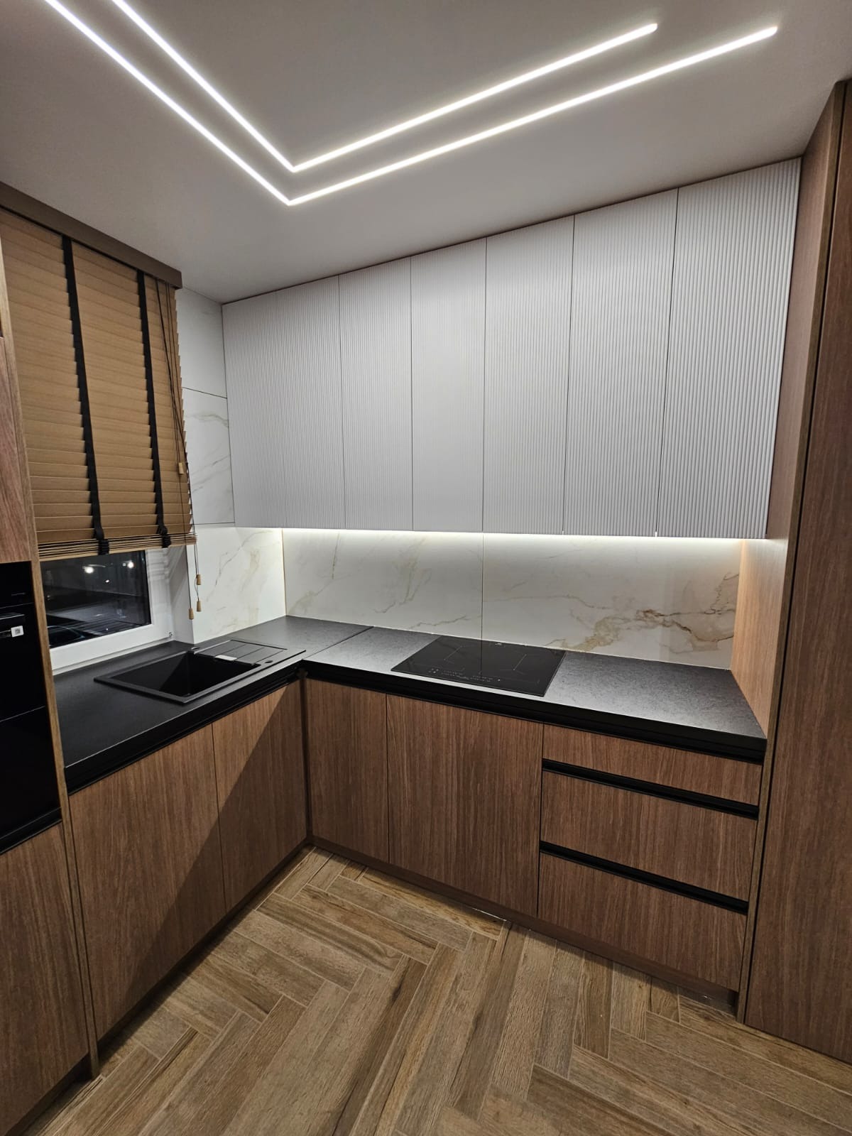 Nowoczesna kuchnia z drewnianymi frontami szafek dolnych, białymi ryflowanymi szafkami górnymi, czarnym blatem i podświetleniem LED podszafkowym oraz sufitowym, z podłogą ułożoną w jodełkę.