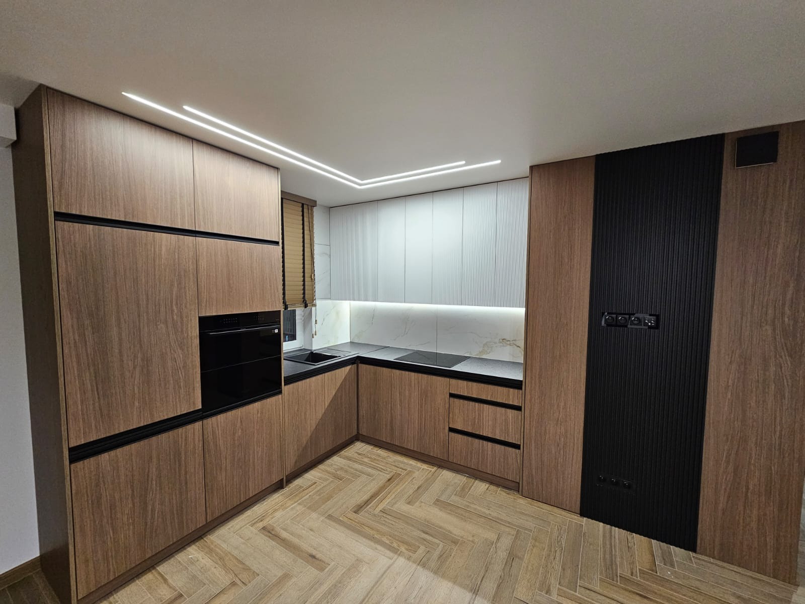 Nowoczesna kuchnia z drewnianymi frontami, czarnymi akcentami i podświetleniem LED, parkiet ułożony w jodełkę.