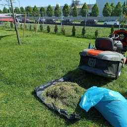 Prace Ogrodowe Michał - Pomarańczowo-szary traktor ogrodowy Husqvarna na trawniku, obok czarna plandeka z pokosem i niebieski worek na śmieci, w tle rząd strzyżonych drzew i budynek przemysłowy.