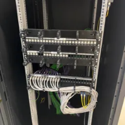 Panel krosowniczy z kablami w szafie rack. Widoczne porty RJ45 i uporządkowane przewody w kolorach: białym, zielonym, fioletowym i żółtym. Estetyczna instalacja.