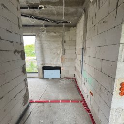 S-PRO Rafał Beryt - Surowy stan budynku z widocznymi instalacjami: czerwone rury przy ścianie, białe kable pod sufitem. Ściany z pustaków, okno w tle. Widoczne puszki elektryczne.