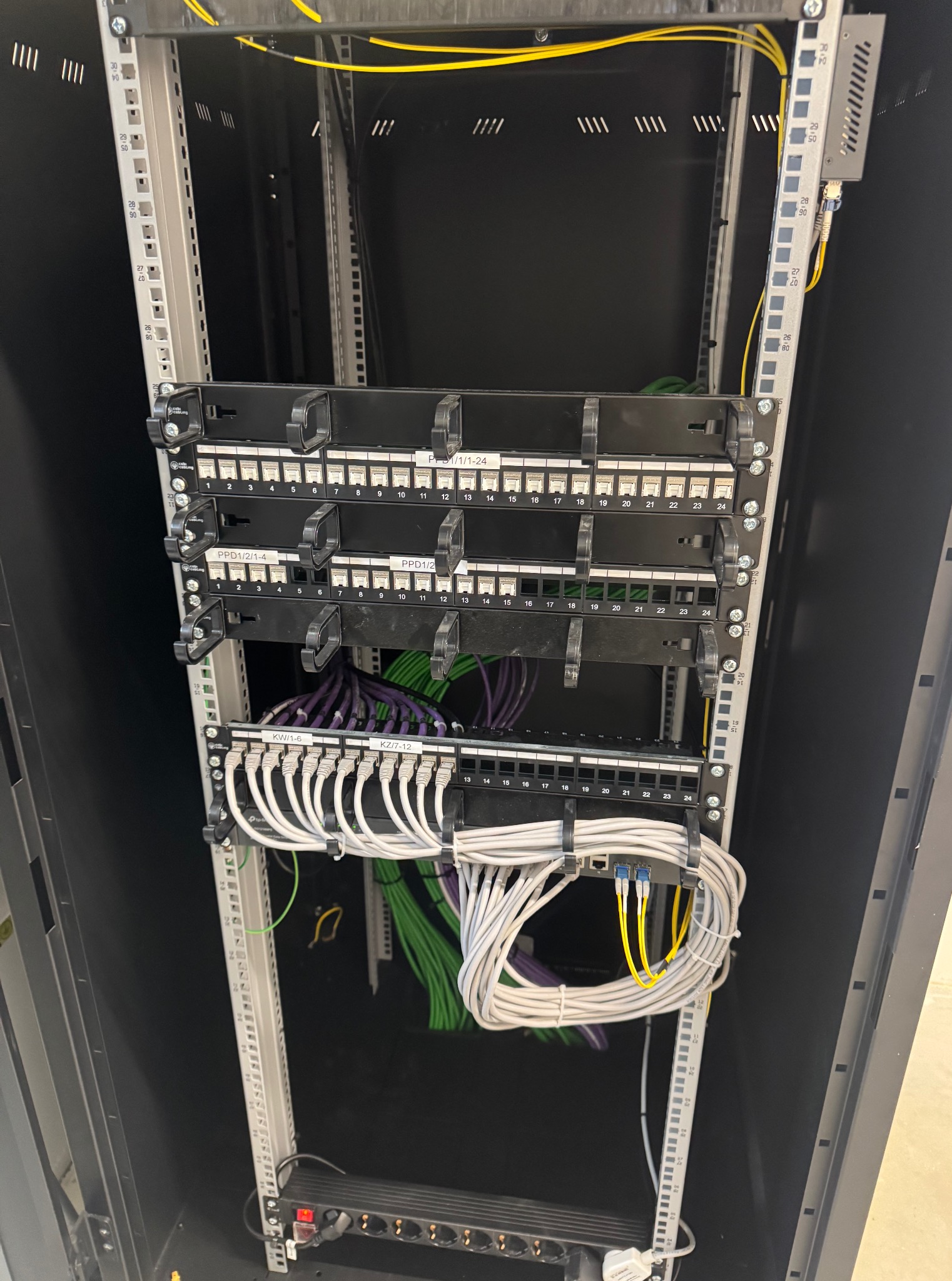 Panel krosowniczy z kablami w szafie rack. Widoczne porty RJ45 i uporządkowane przewody w kolorach: białym, zielonym, fioletowym i żółtym. Estetyczna instalacja.