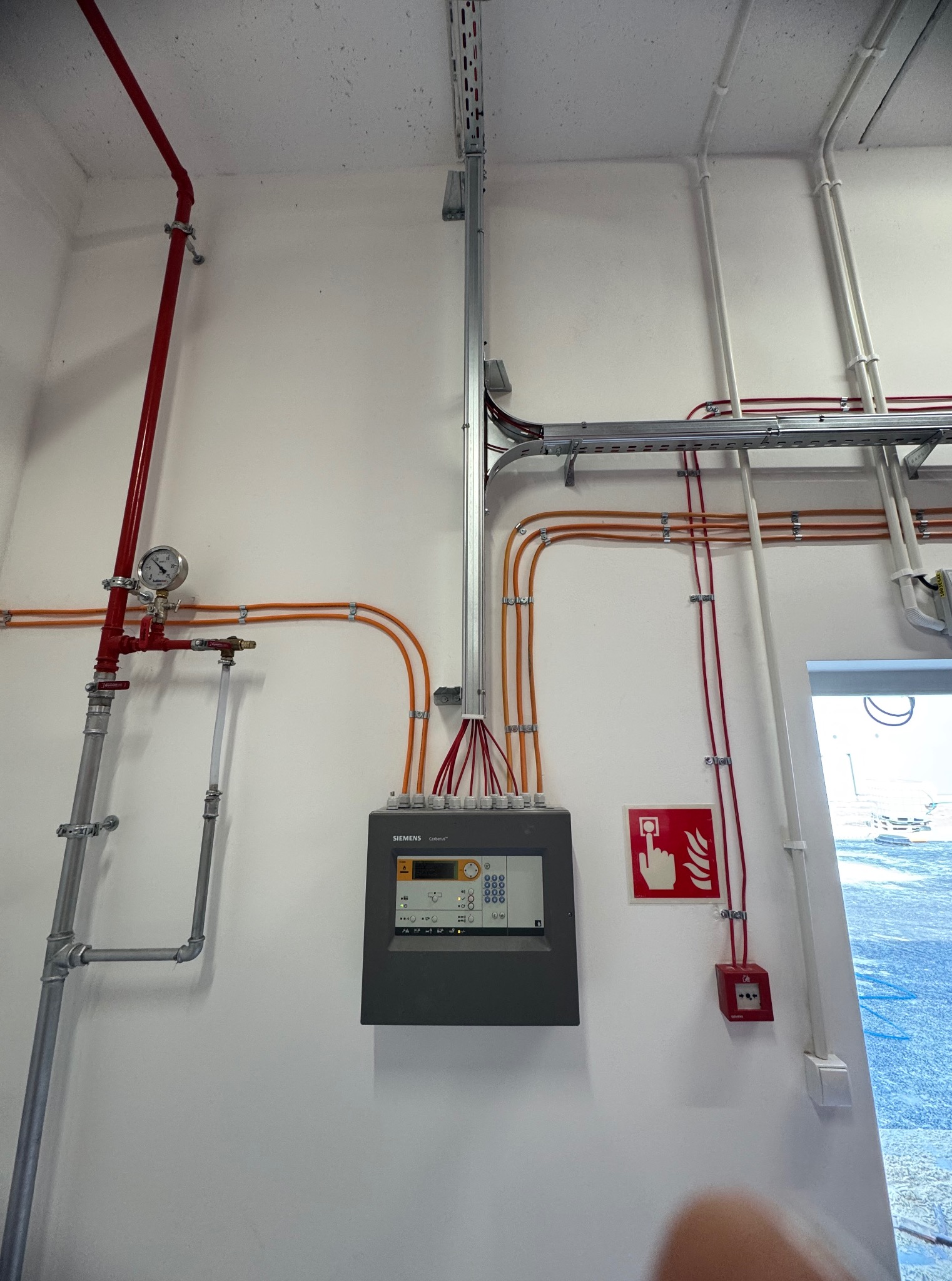 Szara centrala alarmowa Siemens Cerberus na białej ścianie, z widocznymi przewodami, rurami instalacji przeciwpożarowej i przyciskiem alarmowym. Widok z dołu.