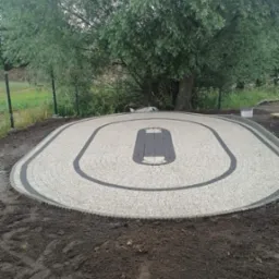 Owalny plac z jasnoszarej kostki brukowej z ciemnoszarą obwódką i centralnym elementem w kształcie toru wyścigowego, otoczony ziemią i zielenią.