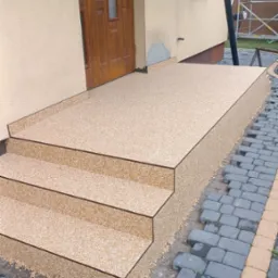Wykończone schody wejściowe do domu pokryte beżową, drobną posadzką kamienną, z widocznymi detalami wykończenia i kostką brukową obok.