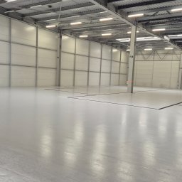 EPOXYPLANET Posadzki żywiczne Kamil Burda - Jasnoszara, gładka posadzka w hali magazynowej z panelowymi ścianami i metalową konstrukcją dachu. Widoczne czarne linie podziału na posadzce i numery na ścianach.